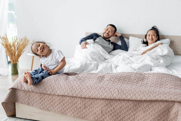 cheerful asian kid sitting on bed near blurred parents - Foto, imagen