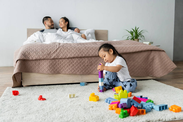 niña jugando bloques de construcción cerca de padres borrosos acostados en la cama en el fondo - Foto, Imagen