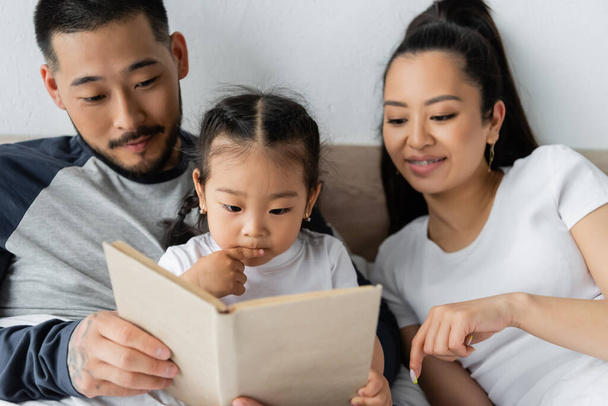 happy asian parents reading book to toddler daughter in bed - Foto, imagen