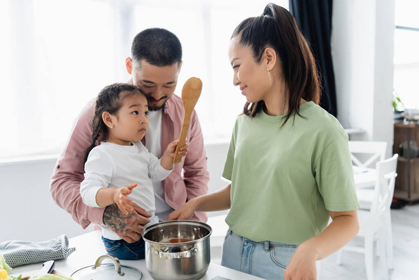 asian mother smiling while looking at daughter near husband in kitchen  - Foto, Bild
