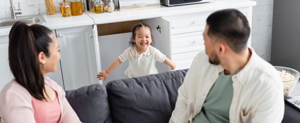 happy asian kid hiding in kitchen cabinet near blurred parents on sofa, banner - Фото, изображение