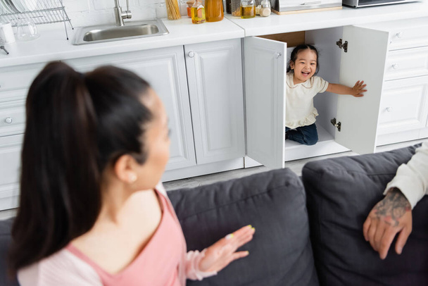 cheerful asian kid hiding in kitchen cabinet near blurred parents on sofa - Foto, imagen