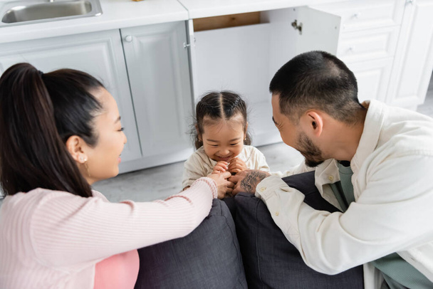 happy asian kid hiding in kitchen cabinet near blurred parents on sofa - Photo, Image