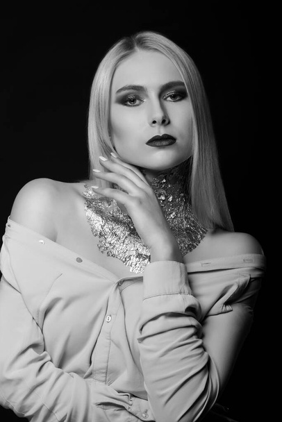 Black and white beauty shoot: fashionable blond model posing at studio with bright makeup and foil on her neck - Foto, imagen