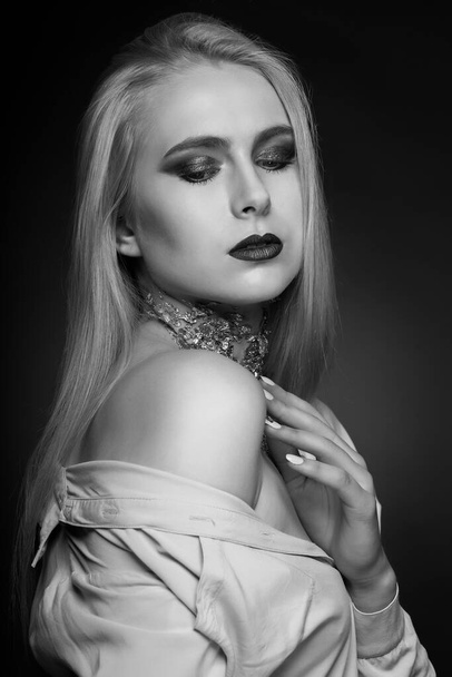 Черно-белая фотосессия: страстная блондинка позирует в студии с ярким макияжем и фольгой на шее - Фото, изображение