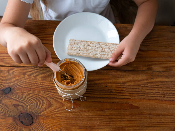 Kind verspreiden pindakaas op knäckebröd eten sandwich houten tafel thuis keuken.School meisje met brood plak volkoren snack.Kid koken ontbijt Biologische voedzame superfood gezonde levensstijl - Foto, afbeelding