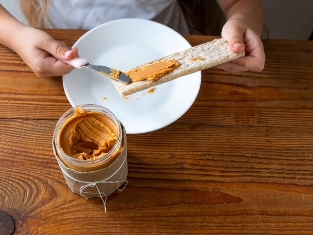 Lapsi levittää maapähkinävoita näkkileipä syö voileipä puinen pöytä kotiin keittiö. Koulun tyttö leipäsiivu täysjyvävälipala. Kid ruoanlaitto aamiainen Orgaaninen ravitseva superruoka terveellistä elämäntapaa - Valokuva, kuva
