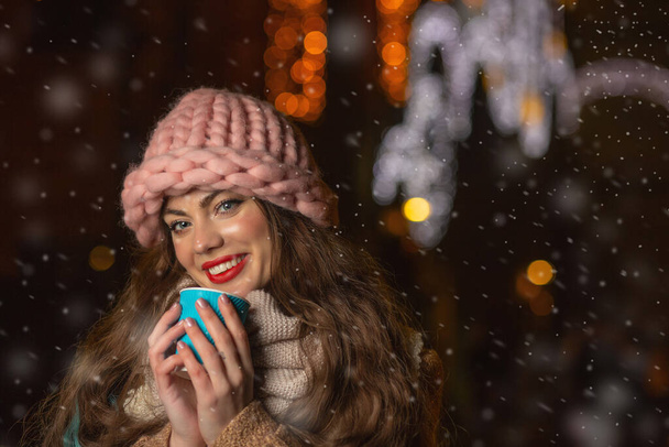 Femme joyeuse porte chapeau tricoté et écharpe boire du café dans la rue en hiver - Photo, image