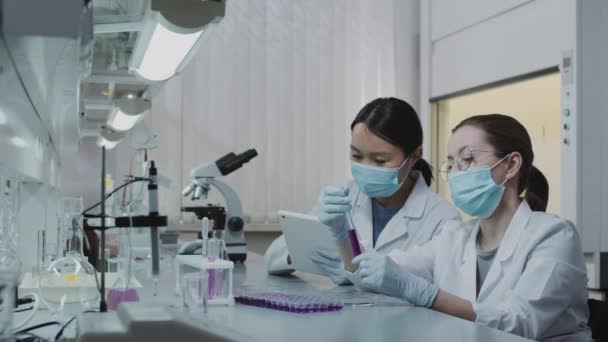 Slowmo PAN kobiet chemików w białych fartuchach i maskach do twarzy omawiających fioletowy roztwór w probówce i notatek na tablecie Mężczyzna Afroamerykanin naukowiec robi badania w tle - Materiał filmowy, wideo