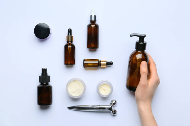 Mão feminina com diferentes produtos cosméticos e ferramenta de massagem facial em fundo leve - Foto, Imagem