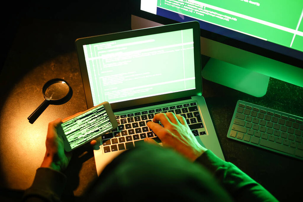 Hacker com telefone celular usando o computador no quarto escuro - Foto, Imagem