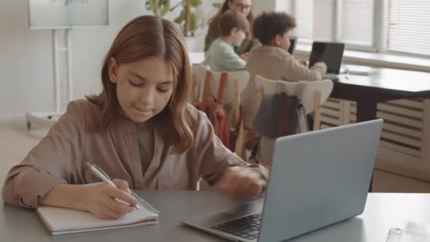 Poitrine-up de jolie fille caucasienne assise au bureau, tapant sur un ordinateur portable et écrivant dans un carnet au premier plan de l'enseignant parlant aux élèves en classe - Séquence, vidéo