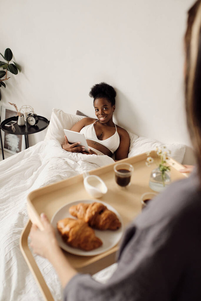 Happy African female looking at her girlfriend holding tray with breakfast - Foto, Bild