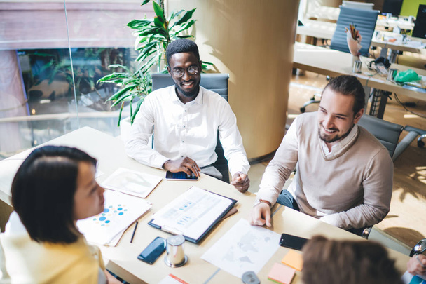 From above of positive group of multiethnic coworkers in casual clothes gathering at table in bright workplace during meeting and discussing marketing strategy together - 写真・画像