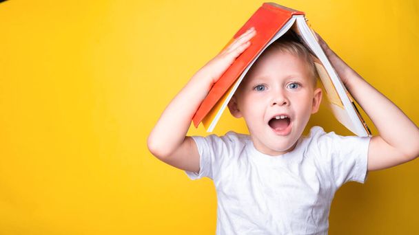 Portrait of funny caucasian blonde school boy with book on his head looking and smiling at camera on yellow background. Back to school and Education concept. - Photo, Image