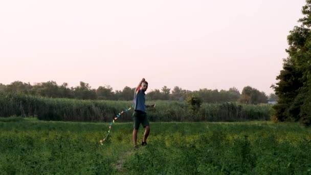 LGBT vlag vlieger. Trage beweging 4k video. Blanke man met dreadlocks staat in het veld en probeert vlieger te vliegen en plezier te hebben in de frisse lucht. Er is niet genoeg wind om te spelen met vlieger in de natuur. - Video