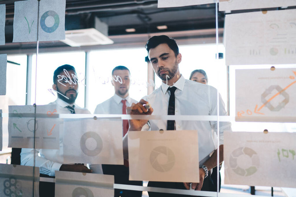 Focused ethnic hipster businessman in formal wear writing on glass wall after thinking new ideas and planning strategy of company with colleagues while standing in modern office - Foto, Imagem