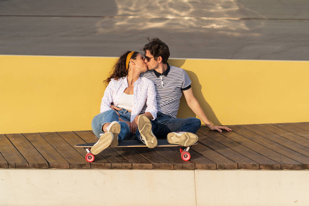 Una coppia di pattinatori alla moda si siede sul longboard baciando. Romantica data di hipster millenari all'aperto - Foto, immagini
