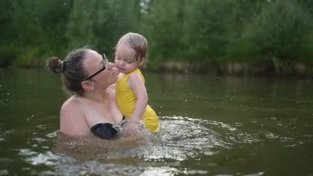 Malá legrační roztomilá blondýnka dítě batole ve žlutém bodysuit smíchu se učí plavat venku u jezera. Plus velikost těla pozitivní usmívající se žena matka dítě plavání v přírodním bazénu. Cákance vody. - Záběry, video