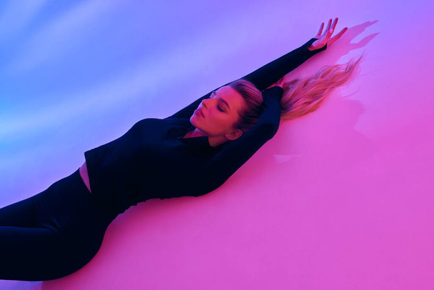 photo shoot of a beautiful girl in a black tracksuit. Against the background of pink light. The girl lies with her hands up - Zdjęcie, obraz