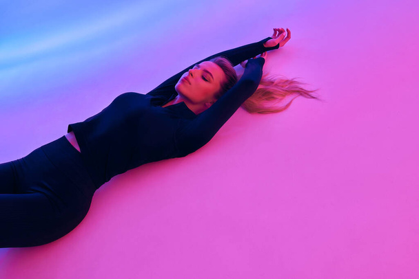 photo shoot of a beautiful girl in a black tracksuit. Against the background of pink light. The girl lies with her hands up - Foto, Bild