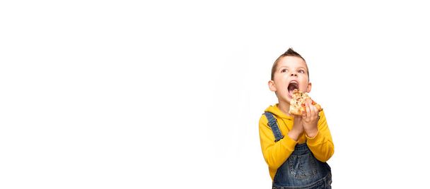 A little cute baby boy in a yellow sweater and denim overalls holds a slice of pizza near his open mouth and looks up in surprise. - 写真・画像