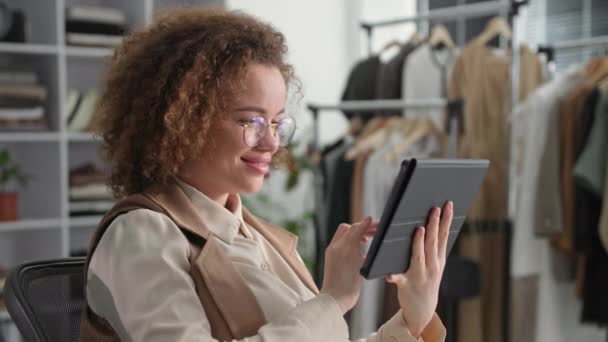 眼鏡の若い女性起業家は、ファッションブティックに座っている間にタブレットを使用して現代的な技術を使用し、オンラインで服を販売しています - 映像、動画