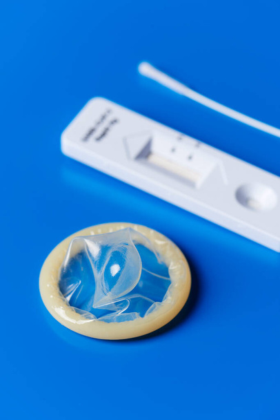 primer plano de un condón enrollado y un dispositivo de prueba de diagnóstico de antígeno covid-19 sobre un fondo azul - Foto, Imagen