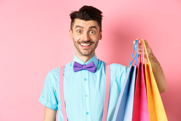 Bild des glücklichen Kerls beim Einkaufen, Papiertüten in der Hand und aufgeregt lächelnd, Käufer mit Rabatten, stehend auf rosa Hintergrund - Foto, Bild