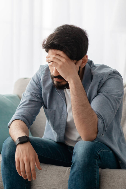 Концепция мужской депрессии. Отчаянный араб трогает лоб, у него проблемы, сидит дома на диване, урожай - Фото, изображение
