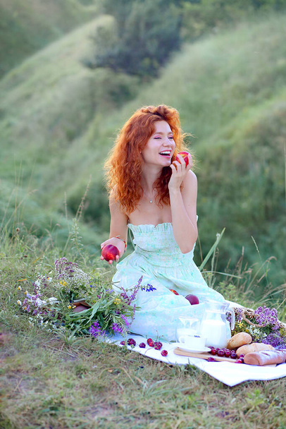 Young and laughing red-haired girl on a picnic while sitting holding in her arms ripe peaches. Food on the table: peaches, cherries, baguettes, milk, herbal tea. Proper nutrition. Harvesting concept - Photo, Image