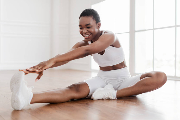 Fröhliche schwarze Frau in weißer Sportbekleidung, die das Bein am Boden ausstreckt - Foto, Bild