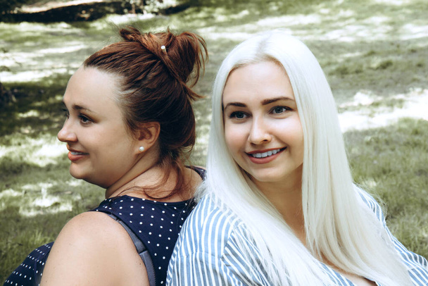 Две молодые улыбающиеся женщины сидят бок о бок в парке - Фото, изображение