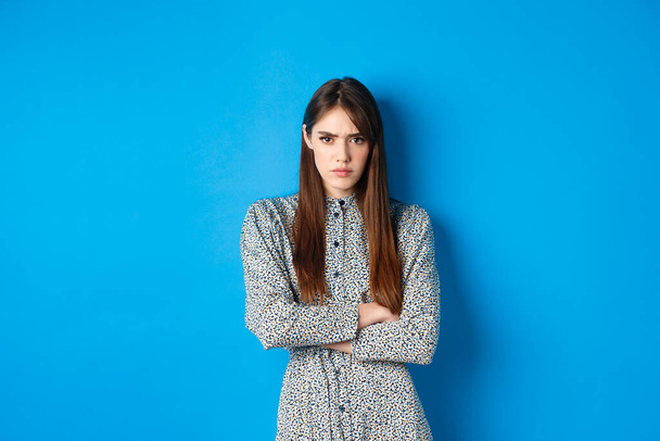 Angry frowning woman in dress, cross arms on chest and feel offended about something unfair, standing upset against blue background - Photo, Image