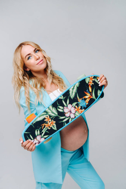 a pregnant girl in turquoise clothes with a skateboard in her hands on a gray background. - Photo, Image
