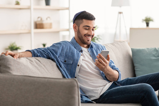 Uomo ebreo sorridente che usa il cellulare seduto sul comodo divano - Foto, immagini