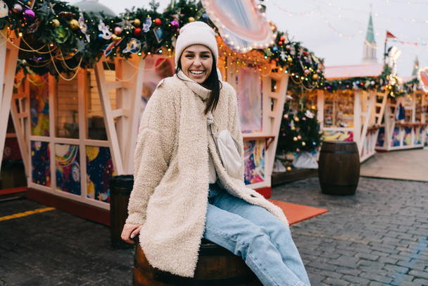 Happy woman in warm wear and hat smiling and looking at camera while sitting on barrel and enjoying Christmas weekend - Fotó, kép