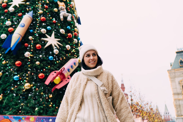 Low angle of happy woman in warm outwear and sweater smiling while standing next to big Christmas tree located in historic city centre - Fotó, kép