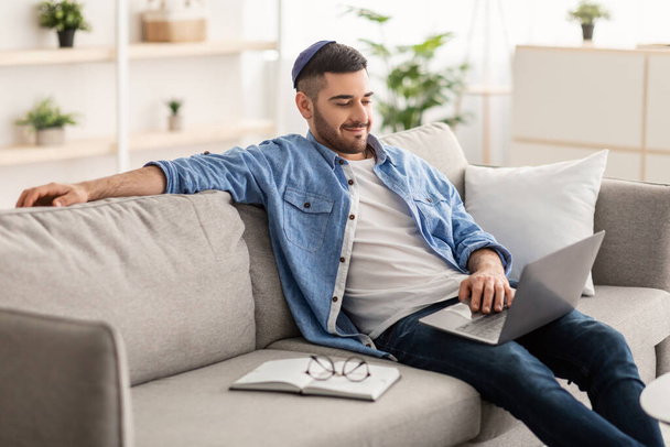 Juif masculin joyeux travaillant sur ordinateur portable à la maison sur le canapé - Photo, image