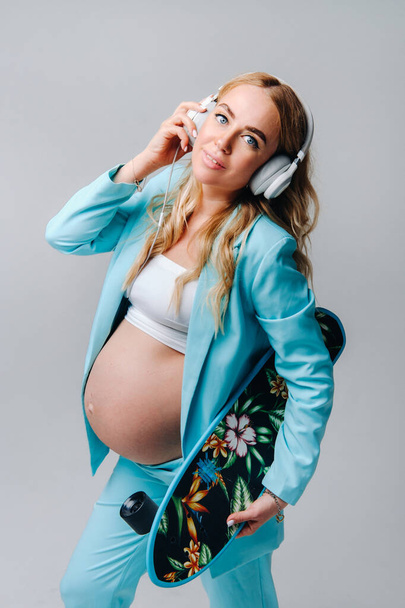 A pregnant girl in a turquoise suit with a skateboard in her hands and headphones stands on a gray background. - Fotó, kép