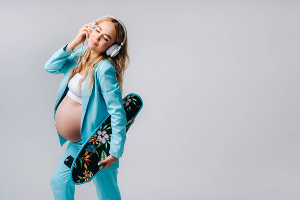 A pregnant girl in a turquoise suit with a skateboard in her hands and headphones stands on a gray background. - Fotografie, Obrázek