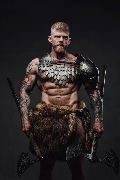 Βίαιος πολεμιστής με τατουάζ φορώντας ελαφριά πανοπλία και γούνα κρατώντας τσεκούρια σε σκοτεινό στούντιο - Φωτογραφία, εικόνα