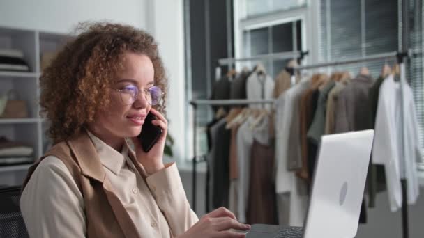 portrait of a young female salesperson talking to a customer on a mobile phone with a customer taking an order in an online store using a laptop in a clothing store - Footage, Video