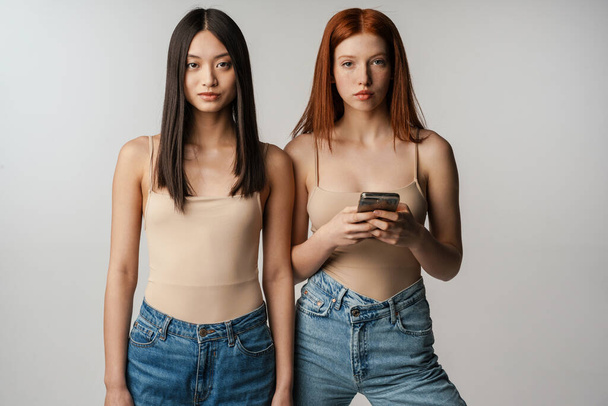 Two young multiethnic women standing posing together over gray wall background, holding mobile phone - Fotografie, Obrázek
