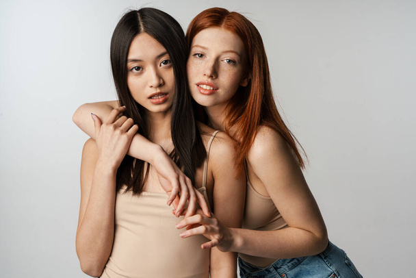 Two young multiethnic women standing posing together over gray wall background, embracing - Foto, Imagem