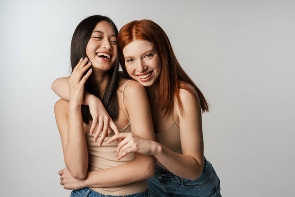 Multiracial two women hugging and laughing at camera isolated over white wall - Foto, afbeelding