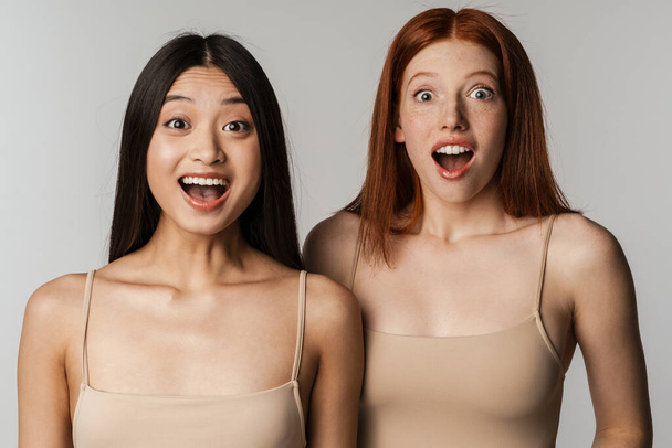 Multiracial two women expressing surprise at camera isolated over white wall - Фото, изображение