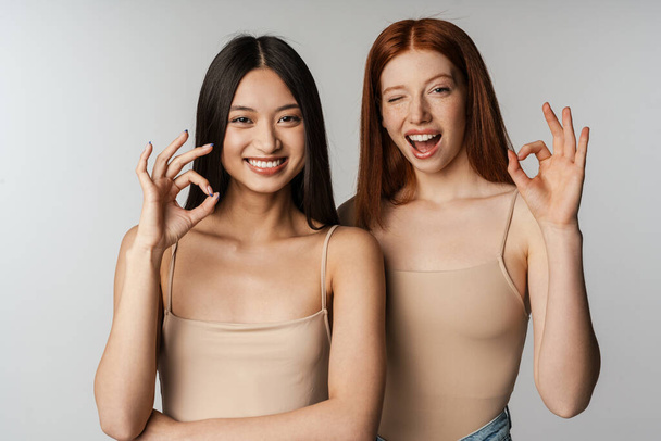 Multiracial two women winking and showing ok sign at camera isolated over white wall - Foto, afbeelding