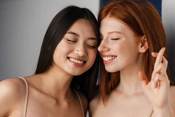 Two smiling young multiethnic women standing together over white wall background, holding fingers crossed for good luck, close up - Foto, Imagen