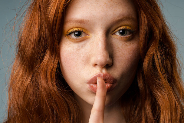 Young ginger woman with makeup showing silence gesture at camera isolated over grey wall - Fotografie, Obrázek